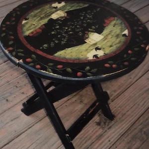 May include: A round, black folding side table with a painted top. The tabletop features a central design of green and white figures on a black background, surrounded by a red border and a floral pattern. The table is supported by black, crossed legs.