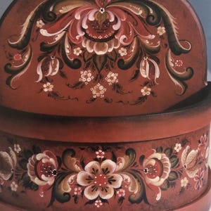 May include: A wooden, hand-painted decorative box with a red background. The box features floral designs in white, pink, and green. The top and front of the box are adorned with intricate floral patterns.