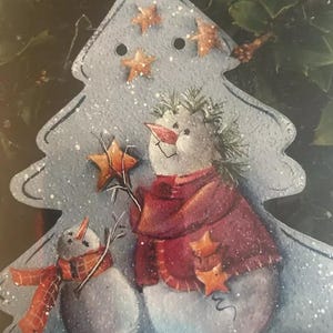 May include: A blue painted wooden Christmas tree with two snowmen. The larger snowman is wearing a red scarf and holding a gold star. The smaller snowman is wearing a red and black plaid scarf. The tree is decorated with gold stars.