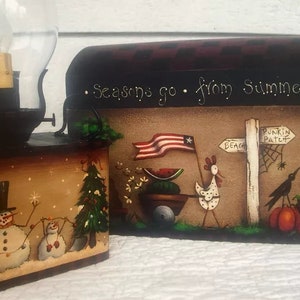 Puede incluir: Una caja de madera decorativa con una lámpara encima, con escenas de temporada pintadas a mano. Un lado representa un muñeco de nieve y un árbol de Navidad, mientras que el otro muestra una escena patriótica con una bandera, una gallina y un espantapájaros. El texto dice "Seasons go from Summer Sun."