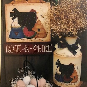 May include: A painted wooden sign with the text "RISE-N-SHINE" and a painted image of a black hen with a blue and white bag, a watermelon slice, and a beehive. A painted milk jug with a black hen design and a bouquet of dried white flowers in a white vase.