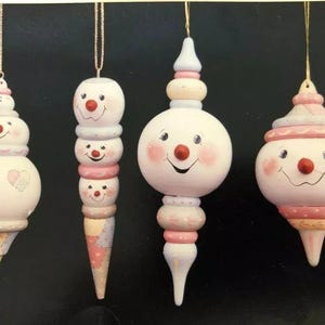 May include: Four snowman-themed Christmas ornaments with smiling faces, rosy cheeks, and red noses. Each ornament is painted white with pink, yellow, and blue accents. They hang from gold-colored strings, perfect for holiday decorating.