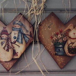 Puede incluir: Dos placas de madera en forma de corazón con temas pintados de invierno y primavera. Una presenta muñecos de nieve, la otra un conejito con una cesta y flores. Ambas tienen colgadores de alambre y detalles de paja.