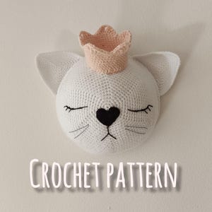 May include: Crochet pattern for a white cat with a pink crown. The cat has black eyes, a black nose, and black whiskers. The text "CROCHET PATTERN" is written below the cat.