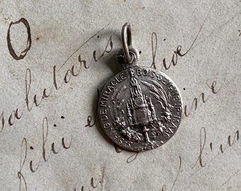 May include: Silver pendant with a detailed image of a church and the text "S.G. DE MIRACLE DU BOIS SEIGNEUR DE GRUPINO" around the edge.