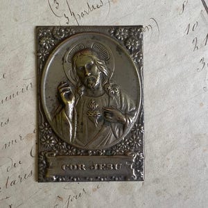 Op de afbeelding: Antieke zilverkleurige religieuze plaquette met een gedetailleerde afbeelding van Jezus Christus in een ovale lijst. De plaquette bevat de inscriptie "COR JESU" en is omgeven door bloemmotieven.