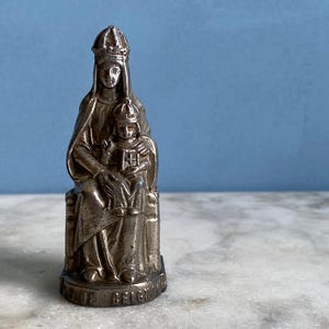 May include: Silver-toned figurine of the Virgin Mary holding a child. The figurine is wearing a crown and a robe. The base of the figurine is inscribed with the words "Our Lady of Belgium".