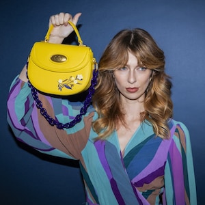 May include: A yellow handbag with a floral design and a purple chain strap. The bag has a gold clasp and is held by a person wearing a colorful patterned shirt.