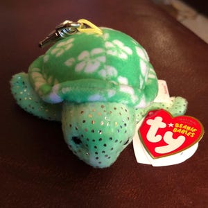 May include: A green plush turtle with white floral patterns on its shell and sparkling accents. It has a metal clip and a red heart-shaped tag that reads "Beanie Babies".