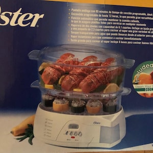 May include: White Oster electric food steamer with a digital timer and two clear glass bowls. The top bowl contains cooked lobster and the bottom bowl contains cooked sushi. The box includes the text "Included Inside" and "Steam Your Cuisine Cookbook".