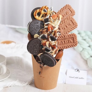 May include: A brown paper cup filled with a chocolate and vanilla dessert topped with chocolate sauce, Oreos, pretzels, and chocolate-covered almonds. The dessert is decorated with chocolate-covered biscuits with the word "LILA" printed on them.