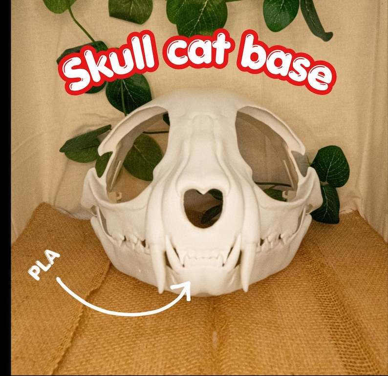 以下が含まれることがあります： 赤色の「Skull cat base」というテキストが入った白い猫の頭蓋骨ベース。 頭蓋骨にはハート型の開口部と目に見える歯があります。 素材はPLAです。