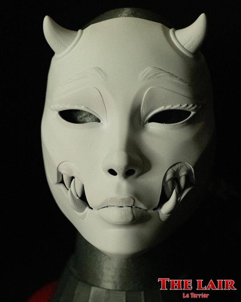 May include: A white mask with a demonic design, featuring curved horns, sharp teeth, and detailed facial features. The mask has a smooth texture and is set against a dark background. The text "THE LAIR Le Terrier" is visible at the bottom.