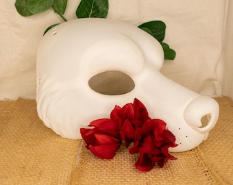 May include: A white, blank animal mask with a long snout and large eye openings. The mask is decorated with two red flowers and green leaves. The mask is sitting on a burlap surface.