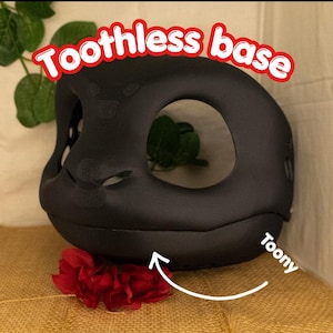 May include: A black "Toothless base" with a large oval opening and small eye holes. The base has a rounded shape and is labeled "Toony". A red flower is at the base of the object.