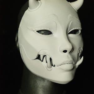 May include: A white mask with a stylized face, featuring curved horns, detailed eye and mouth features, and sharp teeth. The mask has the text "THE LAIR" in red at the bottom.