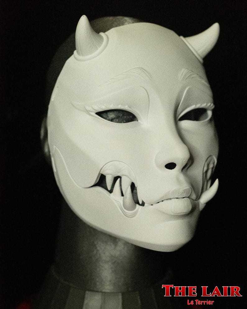 May include: A white mask with a stylized face, featuring curved horns, detailed eye and mouth features, and sharp teeth. The mask has the text "THE LAIR" in red at the bottom.