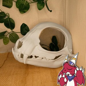 以下が含まれることがあります： 大きな開口部のある白い猫の頭蓋骨の装飾品。植物や小さなオブジェクトを入れることができます。頭蓋骨は詳細な特徴があり、緑の葉のあるニュートラルな背景に配置されています。