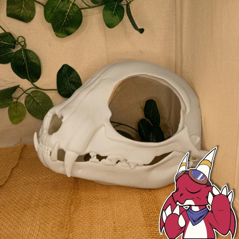 以下が含まれることがあります： 大きな開口部のある白い猫の頭蓋骨の装飾品。植物や小さなオブジェクトを入れることができます。頭蓋骨は詳細な特徴があり、緑の葉のあるニュートラルな背景に配置されています。