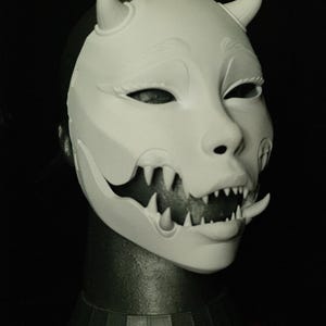 May include: A white mask with a demonic design, featuring horns, sharp teeth, and a stylized face. The mask is displayed on a black background. The text "THE LAIR Le Terrier" is visible at the bottom.