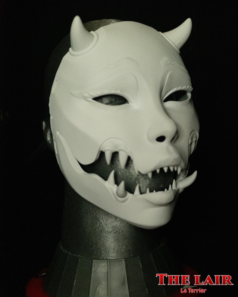 May include: A white mask with a demonic design, featuring horns, sharp teeth, and a stylized face. The mask is displayed on a black background. The text "THE LAIR Le Terrier" is visible at the bottom.