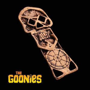 May include: A bronze-coloured metal pendant featuring a skull design at the top and a wheel-like symbol at the bottom. The pendant is rectangular with a cutout at the bottom. The Goonies logo is at the bottom.