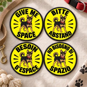 Könnte beinhalten: Vier runde gelbe Aufnäher mit schwarzem Rand, die jeweils einen schwarz-braunen Hund mit rotem Halsband zeigen. Der Text lautet GIVE ME SPACE, BITTE ABSTAND, BESOIN D'ESPACE und HO BISOGNO DI SPAZIO, angeordnet um die Hundeabbildungen.