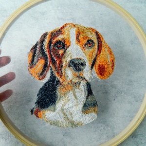 Op de afbeelding: Borduurwerk van een beagle in een houten borduurring. De hond is bruin, zwart en wit met een witte snuit en bruine ogen.