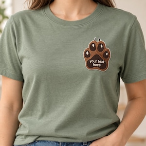 Könnte beinhalten: Ein salbeigrünes T-Shirt mit kurzen Ärmeln und einem gestickten braunen Pfotenabdruck-Aufnäher auf der linken Brust. Der Aufnäher enthält den Text "your text here" in weißer Schrift, zentriert im dunkelbraunen Pfotenballen-Design.