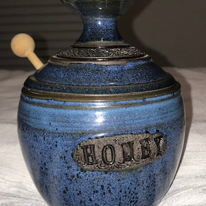 May include: A blue and brown ceramic honey pot with a wooden dipper. The pot has a decorative design and the word "Honey" is written on the front.