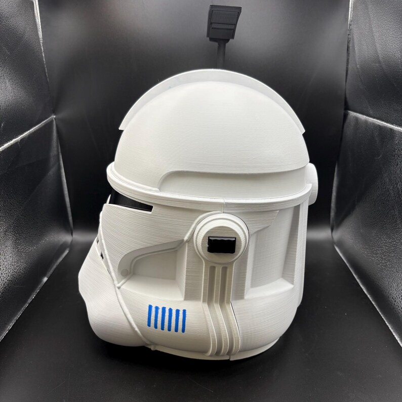 May include: A white, 3D-printed helmet with a distinctive design, featuring a rounded top and angular details. It has a black rectangular visor and a series of blue vertical lines on the side. A black antenna-like structure extends from the top.