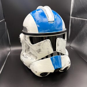 Puede incluir: Casco blanco y azul con visera negra. El casco tiene un aspecto envejecido y desgastado con rayas y detalles azules. El casco es una réplica de un casco de soldado clon.