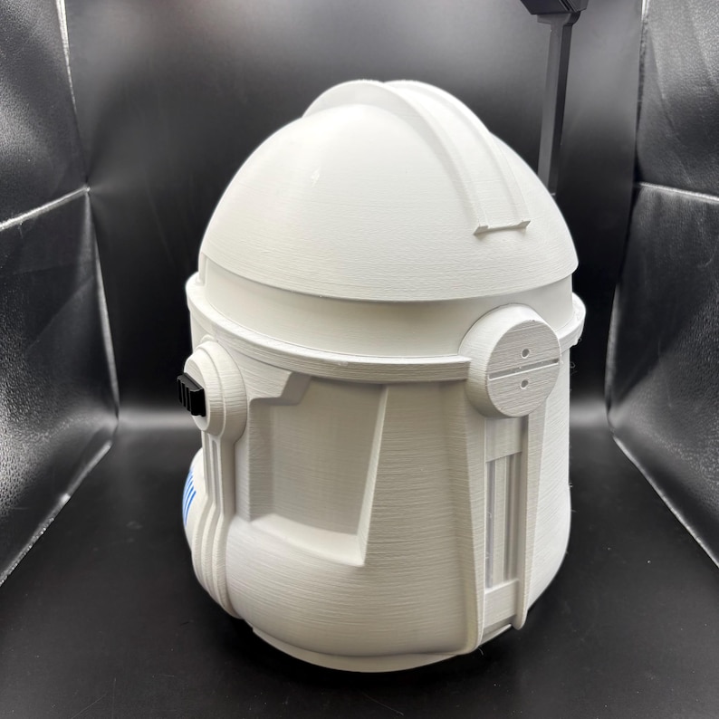 May include: A white, 3D-printed helmet with a rounded top and detailed side panels. The helmet features a visor area and a textured surface, with a few blue stripes visible on the side. The design is reminiscent of a science fiction or military style.