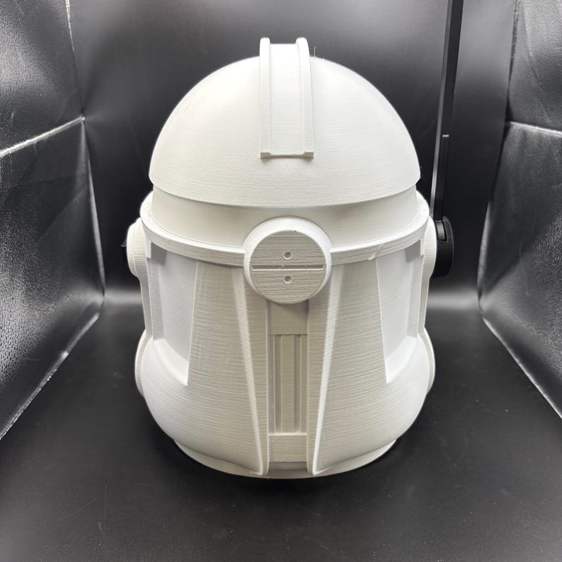 May include: A white, 3D-printed helmet with a rounded top and detailed front panel. The helmet features a visor-like structure and circular side details. The overall design suggests a futuristic or sci-fi theme, possibly for cosplay or display.