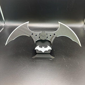 May include: A black and silver batarang, a stylized weapon with a bat-shaped design. The batarang has a central circular emblem and a bat symbol on the base. The edges are silver, and the overall design is symmetrical.