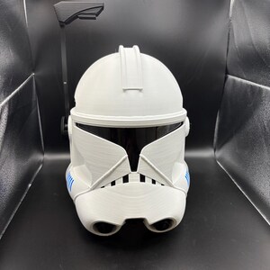 May include: A white, textured helmet with a black visor and blue detailing. The helmet features a unique design with a central ridge and a black attachment extending upwards. The design is reminiscent of science fiction or fantasy themes, popular in film and television.