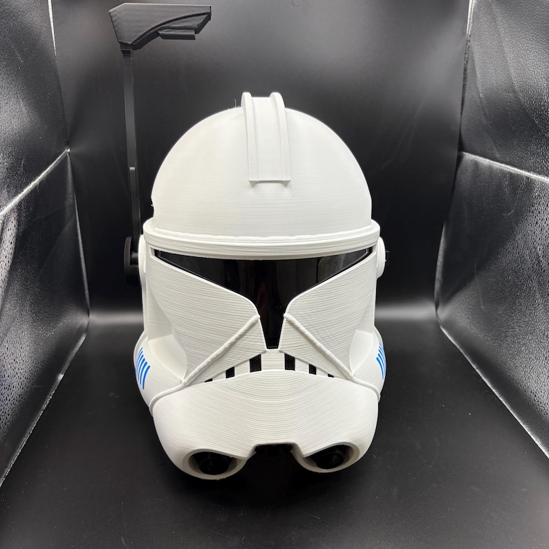 May include: A white, textured helmet with a black visor and blue detailing. The helmet features a unique design with a central ridge and a black attachment extending upwards. The design is reminiscent of science fiction or fantasy themes, popular in film and television.