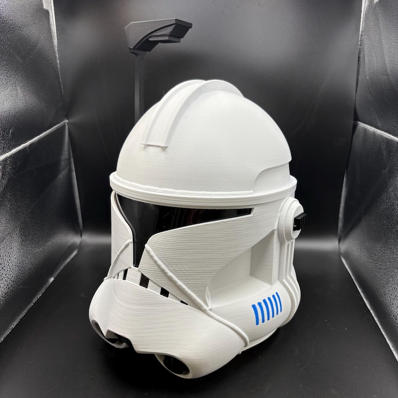 May include: A white, 3D-printed helmet with a black visor and blue detailing. The helmet features a detailed design with a rounded top and a chin piece. A black, rectangular piece is attached to the top of the helmet.