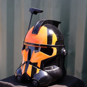 Puede incluir: Un casco negro y naranja con visera y antena. El casco tiene un acabado brillante y presenta rayas y formas geométricas naranjas y negras. El casco es probablemente un disfraz o un objeto de colección.