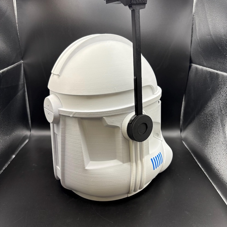 May include: A white, 3D-printed helmet with a black visor mount. The helmet has a detailed design with ridges and a blue stripe detail on the side. The helmet is a replica of a clone trooper helmet.