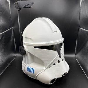 May include: White Clone Trooper helmet with a black visor and antenna. The helmet features blue markings and a detailed design. The helmet is made of a textured material, reminiscent of a Star Wars Clone Trooper.