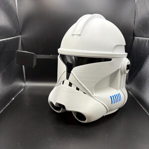 May include: White and black helmet with a visor and a black rectangular attachment. The helmet has blue accents and a detailed design, likely for cosplay or display. The helmet is made of a hard material.