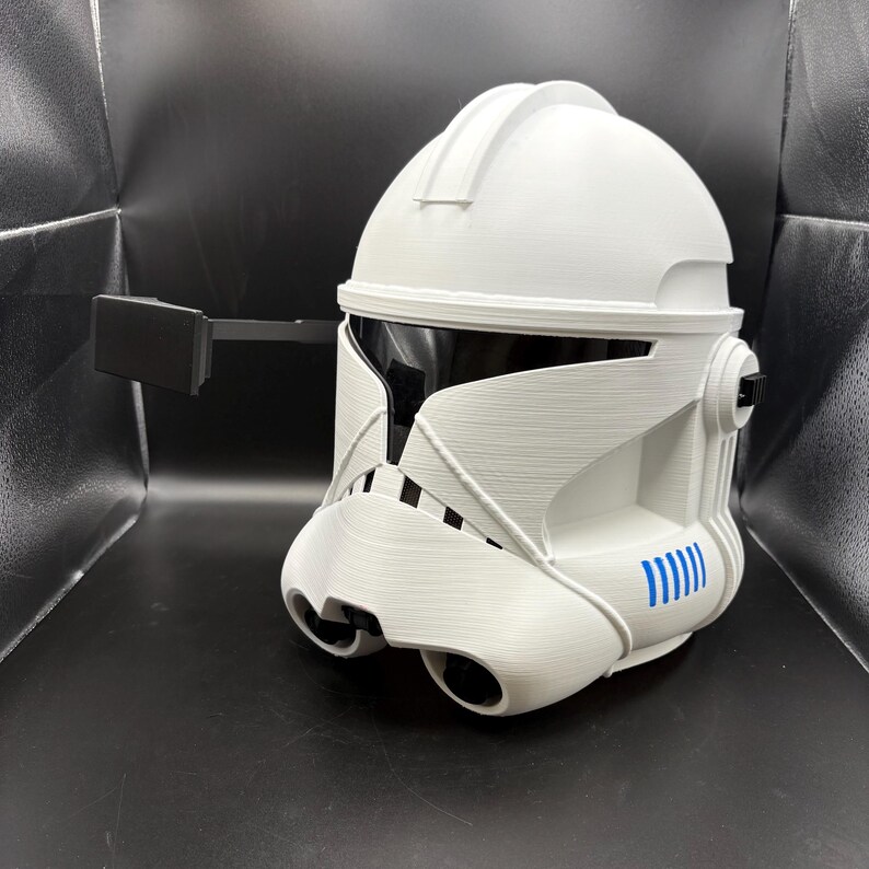 May include: White and black helmet with a visor and a black rectangular attachment. The helmet has blue accents and a detailed design, likely for cosplay or display. The helmet is made of a hard material.