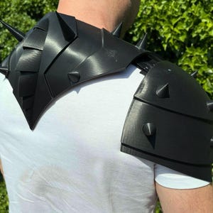 May include: Black shoulder armor with multiple conical spikes. The armor is made of a rigid material and is worn over a white shirt. The design features layered plates and a textured surface, suggesting a fantasy or cosplay theme.