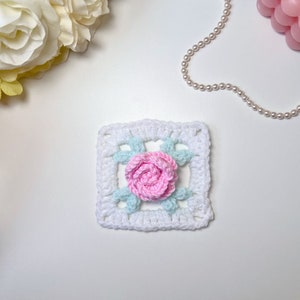 May include: A white crochet square with a pink rose and green leaves in the center.
