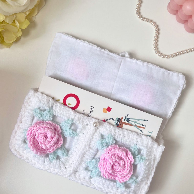 May include: White crocheted book cover with a light blue and pink floral design. The cover is open to reveal a white fabric lining and a book with the text "The Mind".