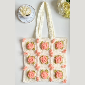 Puede incluir: Una bolsa de mano de crochet blanca con un diseño floral. La bolsa tiene forma cuadrada y presenta un patrón repetido de rosas rosas con hojas verdes. La bolsa tiene dos asas blancas.
