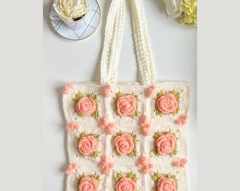 May include: A white crocheted tote bag with a floral design. The bag has a square shape and features a repeating pattern of pink roses with green leaves. The bag has two white straps.