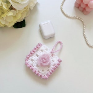 May include: A white crocheted square with a pink flower in the center. The square has a pink border and a pink loop for hanging. The square is on a white surface.