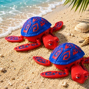 May include: Two red and blue toy turtles with patterned shells and flippers are on a sandy beach. The turtles have a cartoon design, with a blue shell and red body. A seashell and starfish are also visible.